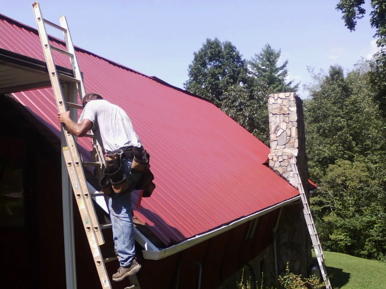 Roofer on a ladder beside a red metal roof and stone chimney — Soffit & Fascia Repair by Roofing Brothers in Duncan Falls
